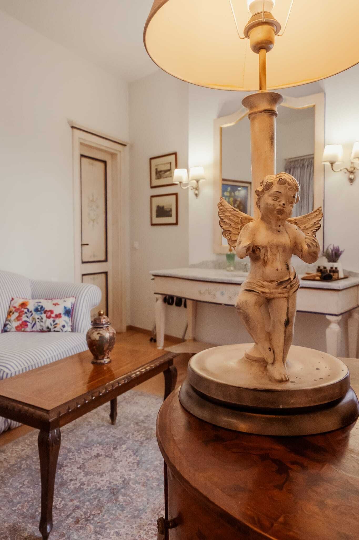 A room with a wooden table, a sofa with a floral cushion, a statue on a pedestal, a mirror, and wall-mounted sconces.