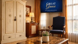 A room with a large wardrobe, a blue armchair, a marble table, a TV screen with 'GREAT GREECE' text, and a window with curtains. - Great Greece Le Convivial Suites & Spa (Xylokastro-Evrostina)