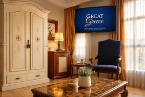 Huoneessa on suuri vaatekaappi, sininen nojatuoli, marmoripöytä, televisioruutu, jossa näkyy teksti ”GREAT GREECE”, ja ikkuna, jossa on verhot.