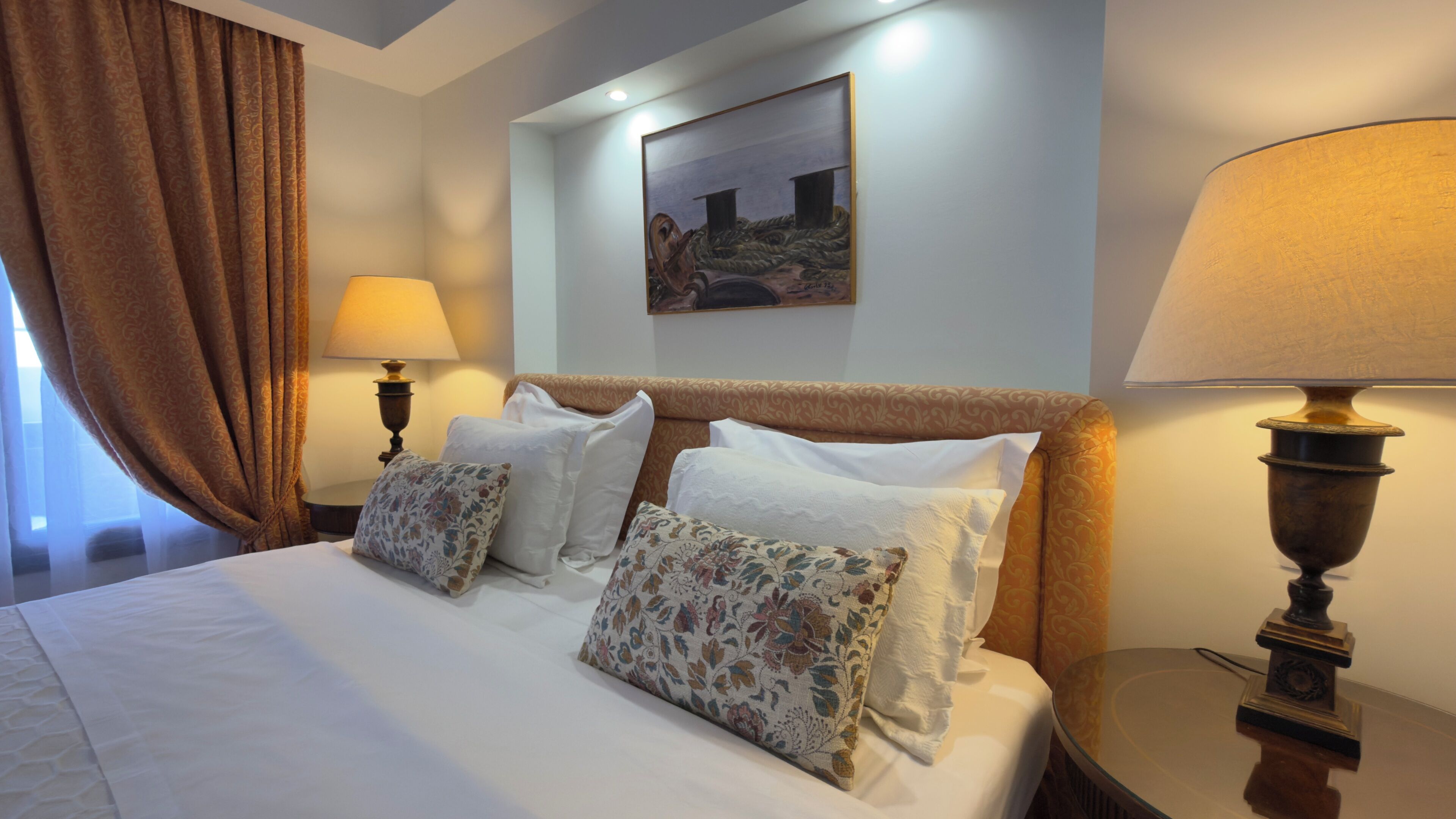 A hotel room with a bed, two pillows, a painting on the wall, and a lamp on the nightstand.