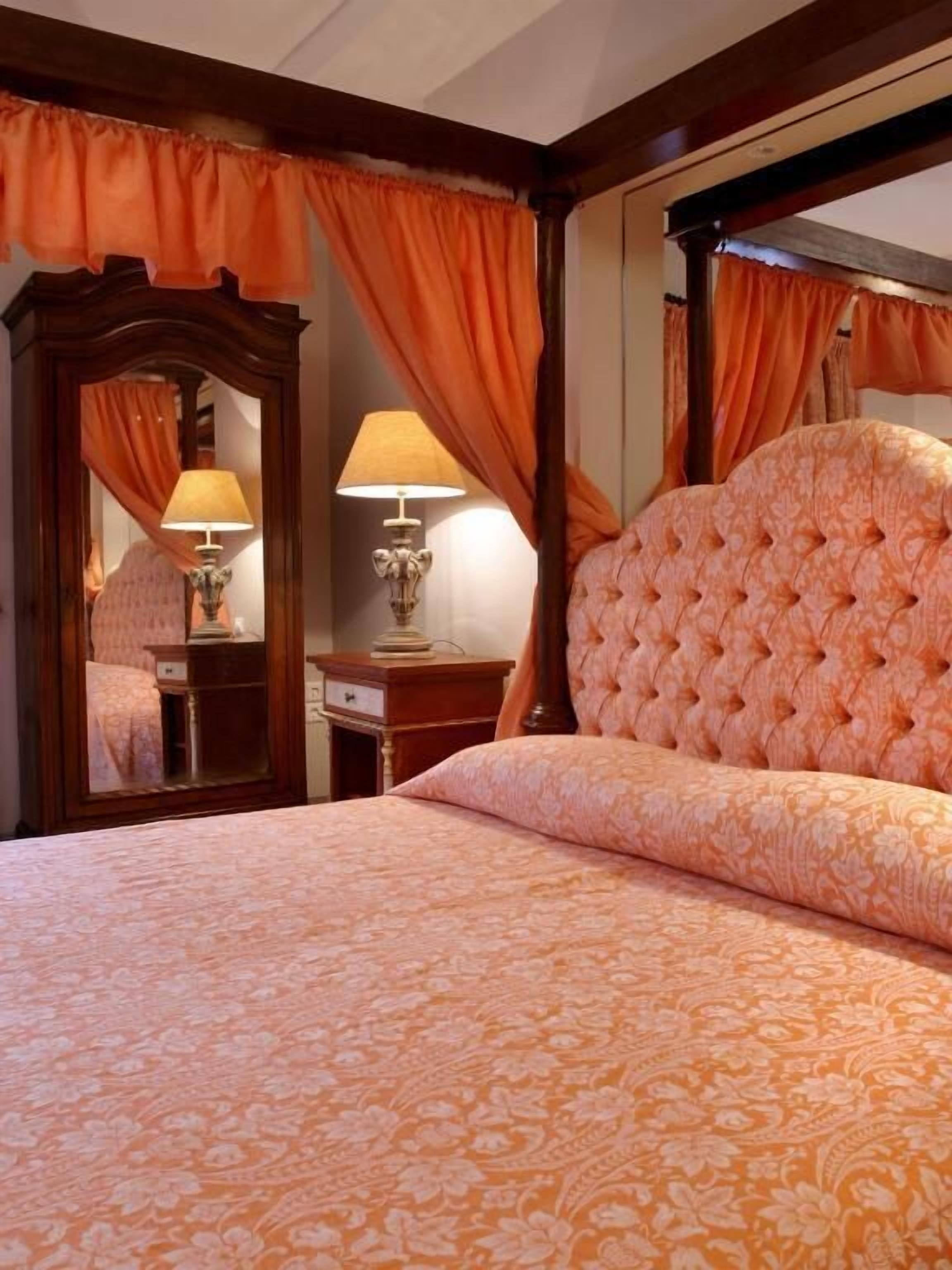 Un dormitorio con una cama grande, un espejo, dos mesitas de noche con lámparas y cortinas anaranjadas.
