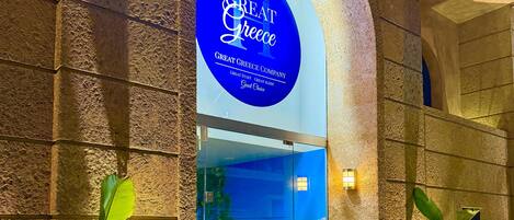 A hotel entrance with a prominent sign reading 'GREAT GREECE COMPANY' and a glass door with a scenic view of a beach.