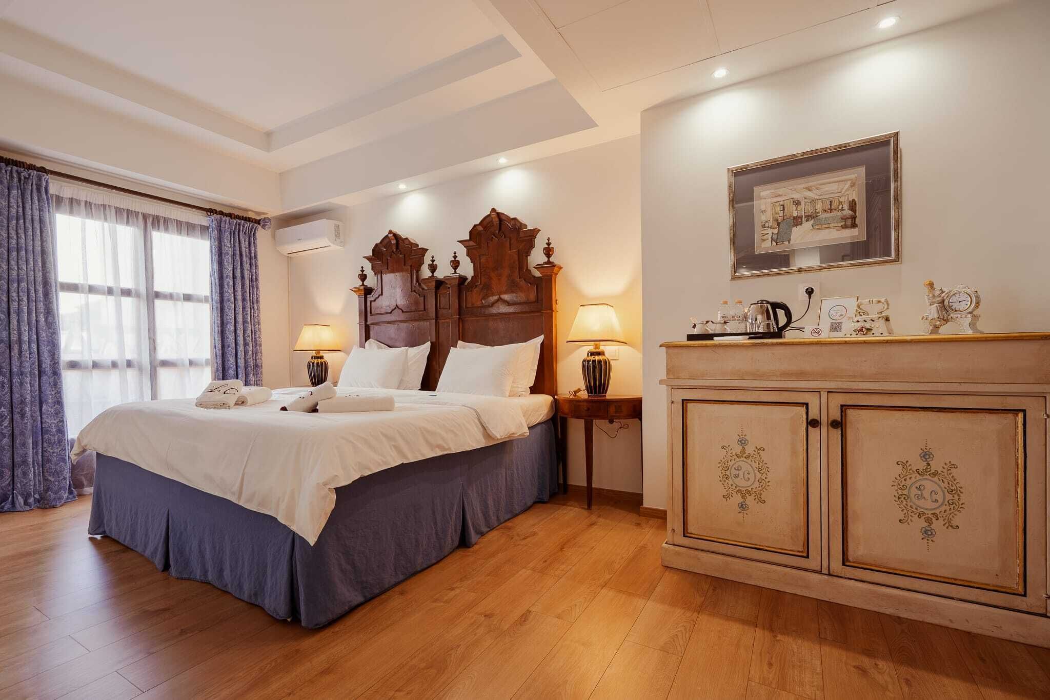 A hotel room with a large bed, a wooden headboard, a nightstand with a lamp, a dresser with decorative items, and a framed picture on the wall.