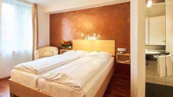 A hotel room with a double bed, a nightstand with a lamp, a small table with a vase of flowers, and a bathroom with a sink and mirror.