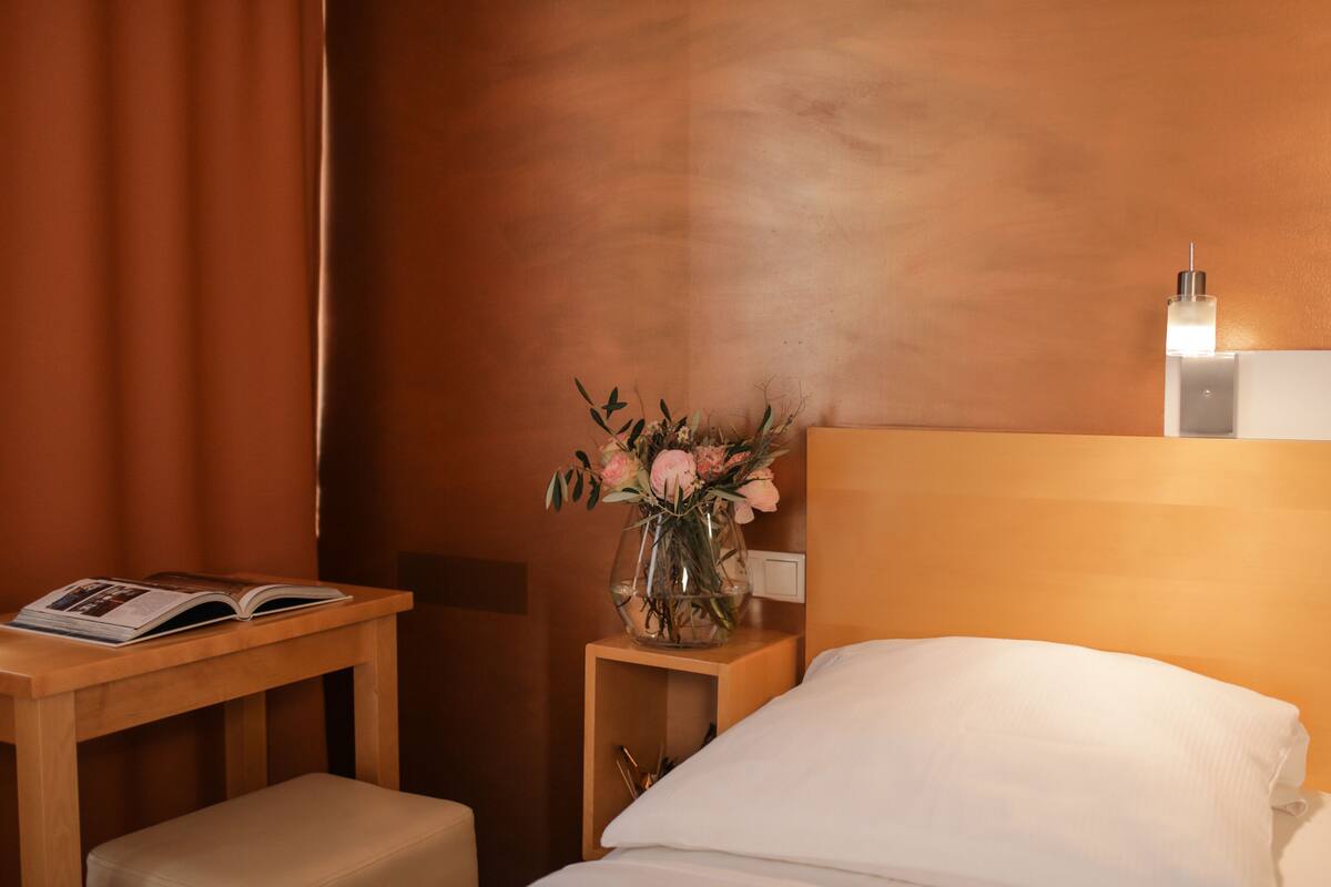 Ein Hotelzimmer mit einem Holzkopfteil, einem Nachttisch mit einem Buch, einer Vase mit Blumen und einem Beistelltisch mit einer Lampe.