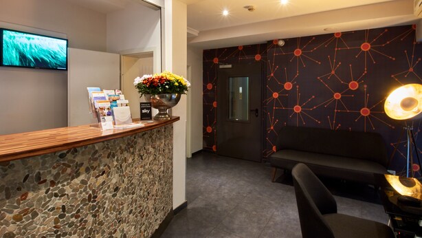 Un área de recepción con mostrador de piedra, televisor, arreglo floral y zona de estar con pared estampada y una lámpara.