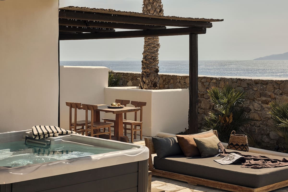A hot tub, wooden furniture, and a stone wall with a view of the sea.