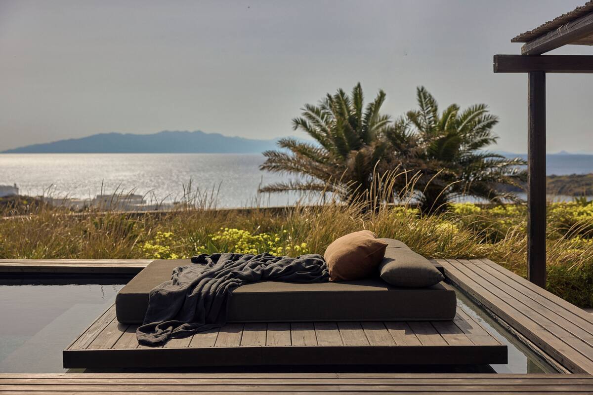 A wooden deck with a mattress and pillows, overlooking a body of water and palm trees.