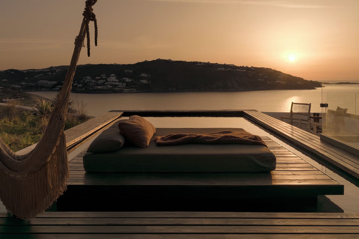 A hammock, a lounge chair, and a wooden deck overlooking a body of water with a sunset.