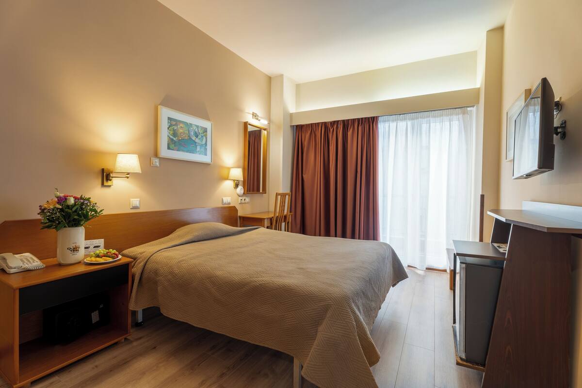 A hotel room with a bed, bedside table with a lamp, a TV, and a desk with a chair.