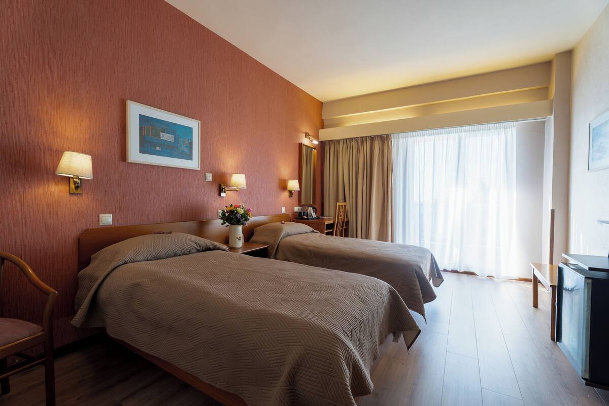 A hotel room with two beds, a desk, a chair, a lamp, and a window with curtains.