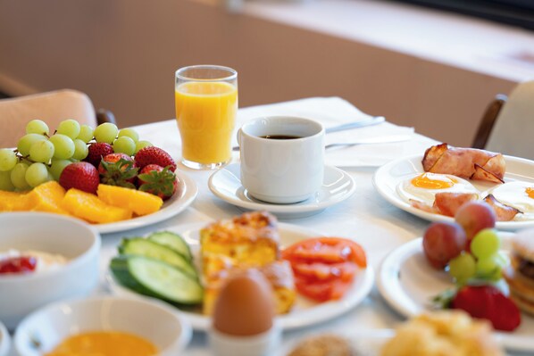 Una mesa de desayuno con diversas frutas, un vaso de jugo de naranja, una taza de café y platos con huevos y tocino.
