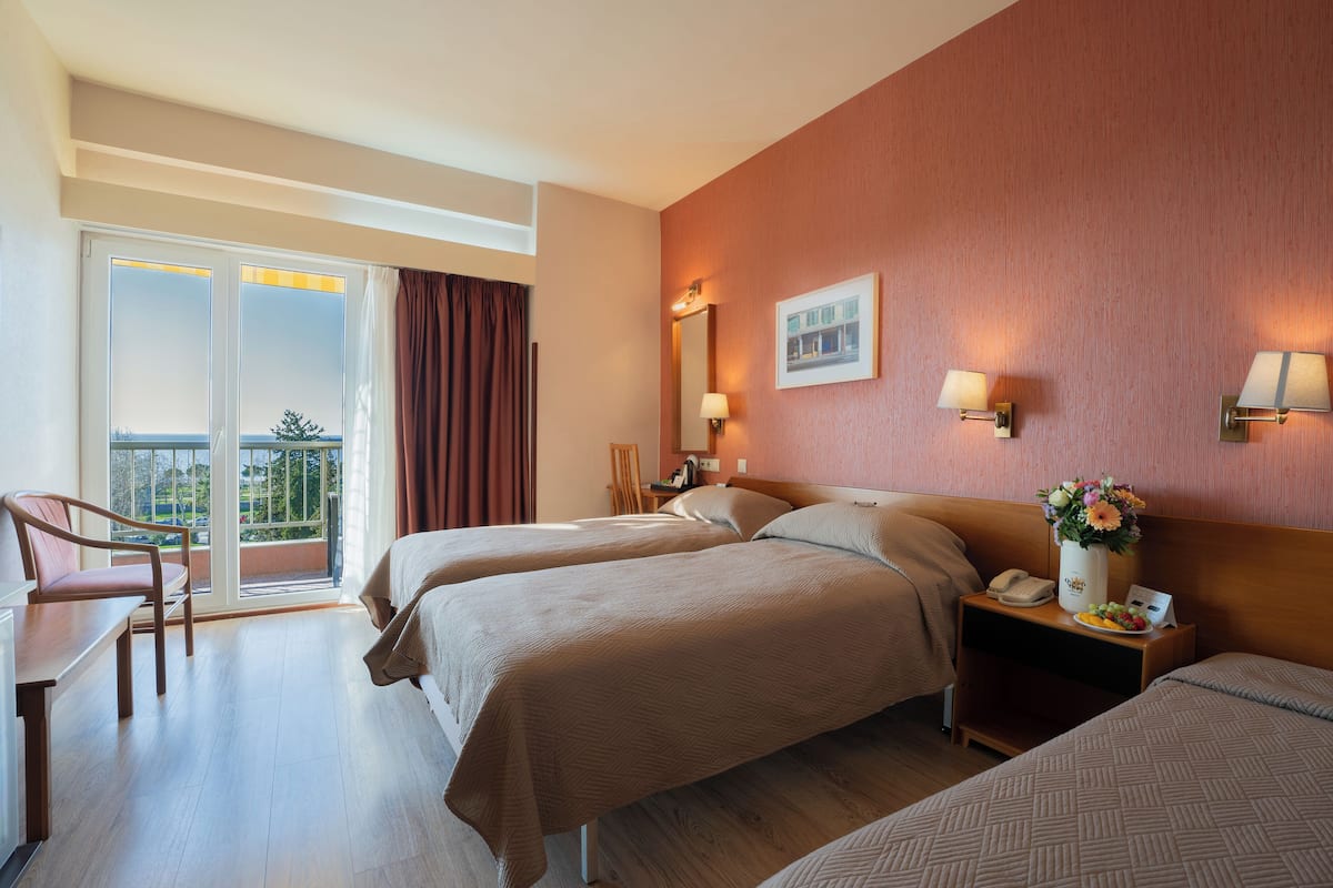 A hotel room with two beds, a desk, a chair, a nightstand with a lamp, and a view of the outdoors.