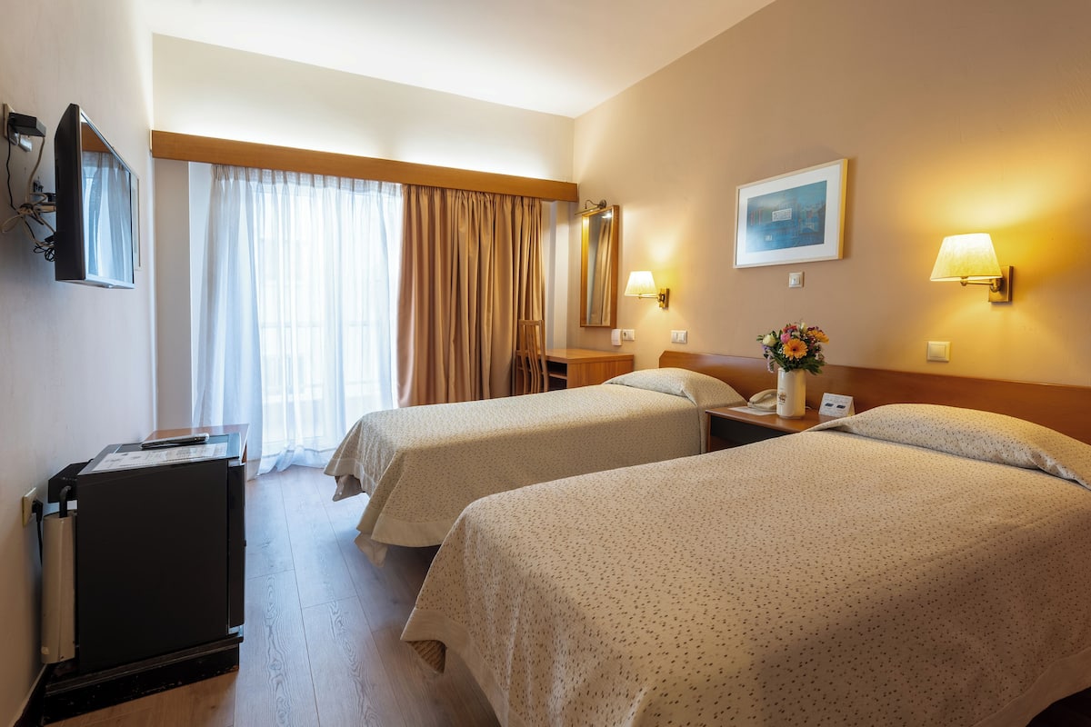 A hotel room with two beds, a desk, a chair, a desk lamp, a telephone, and a small cabinet.