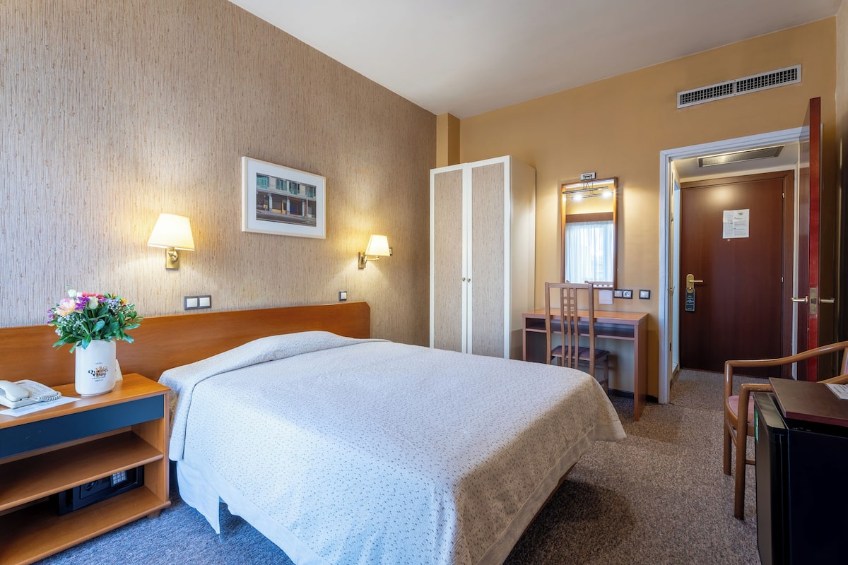 A hotel room with a bed, bedside table, lamp, and a desk with a chair.