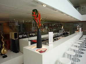 A modern bar area with a white counter, a large vase with red and green leaves, and a glass display of bottles. - Le Fenix Sukhumvit (Bangkok)