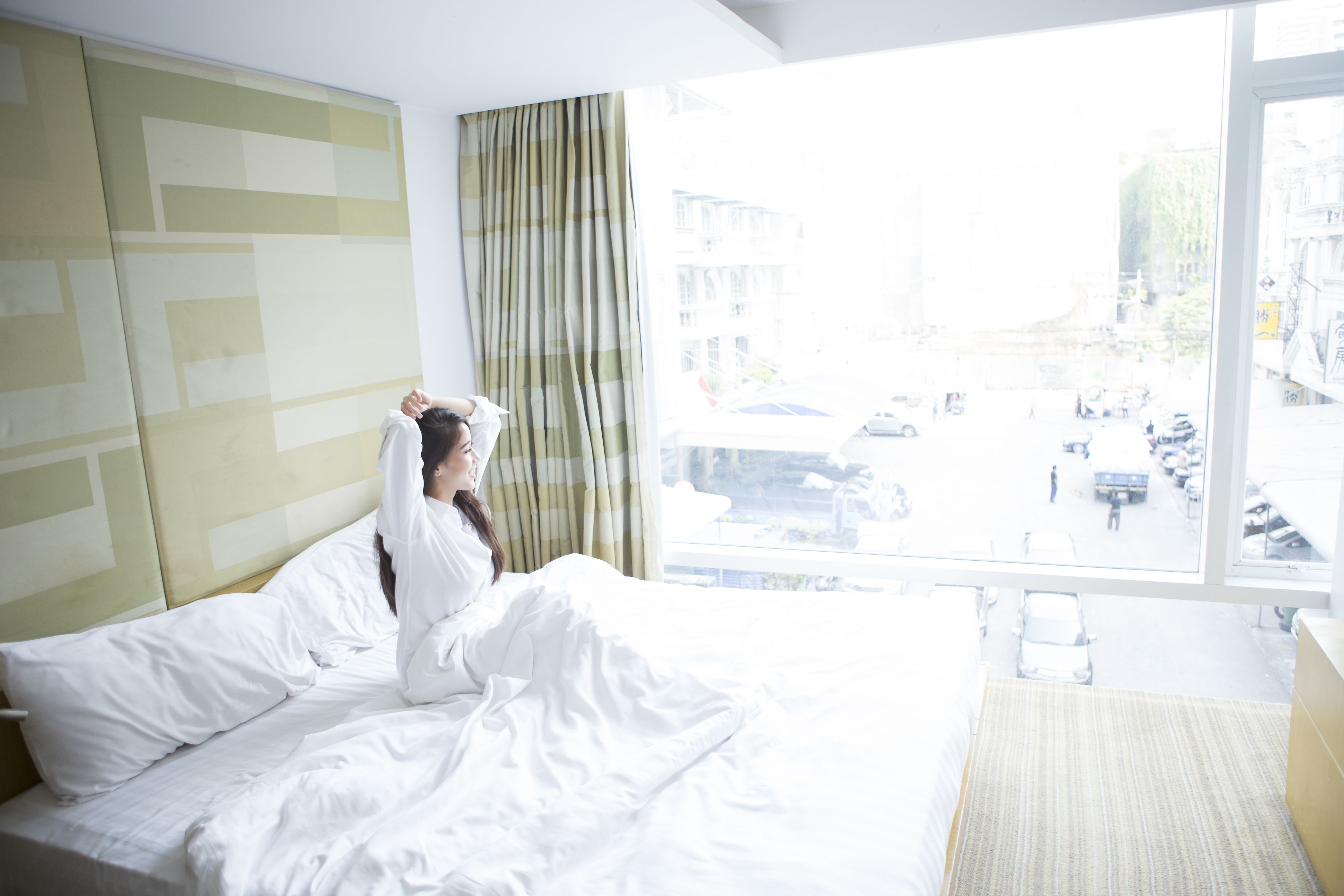 Een hotelkamer met een groot raam, een bed met witte lakens en uitzicht op een levendige straat.
