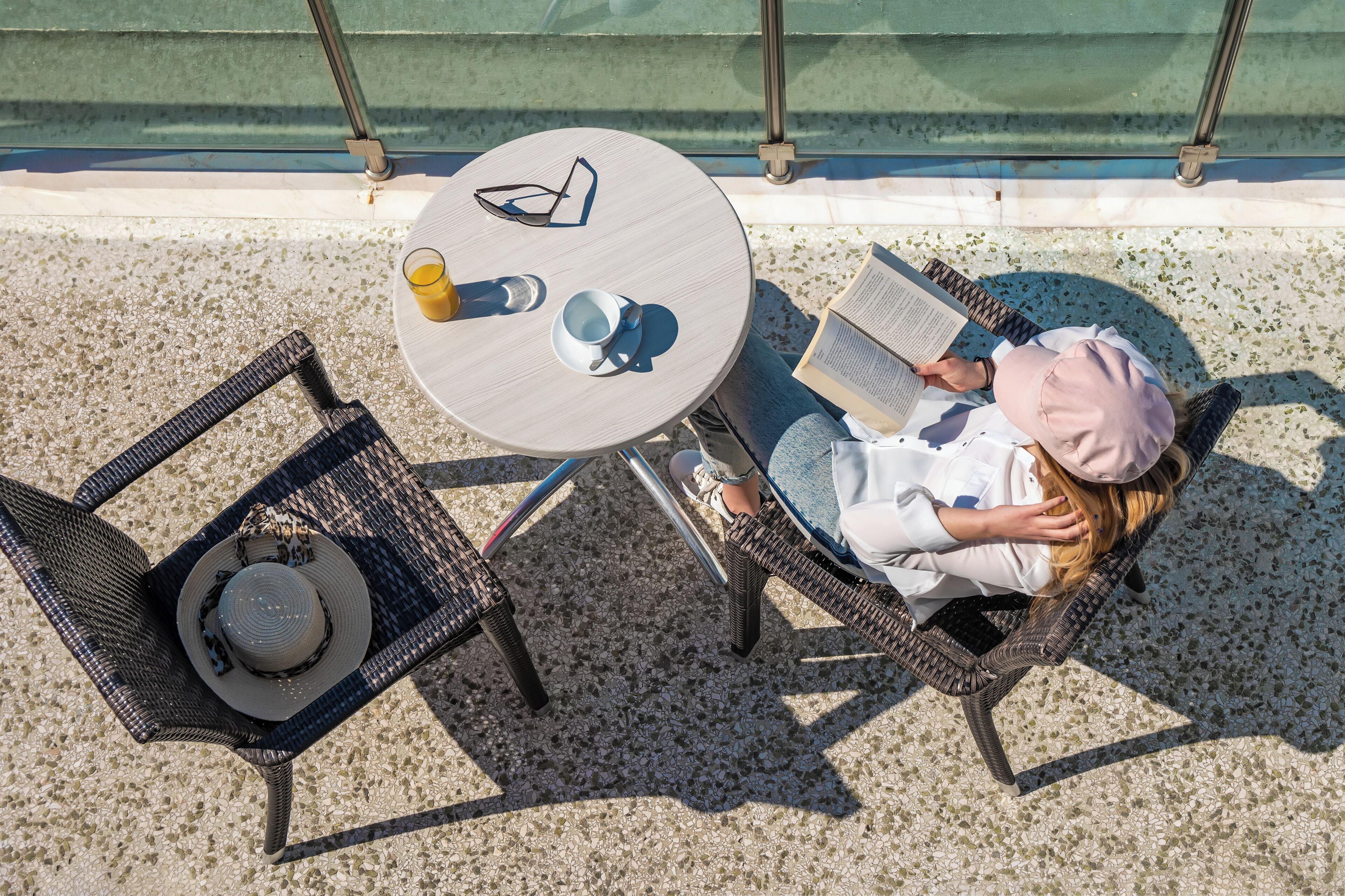 A person reading a book at a round table with a chair, a hat, and a cup on the table.