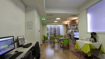 Une chambre d’hôtel équipée d’un bureau, d’un ordinateur, de chaises vertes, d’une table recouverte d’une nappe blanche et d’une fenêtre donnant sur un immeuble.