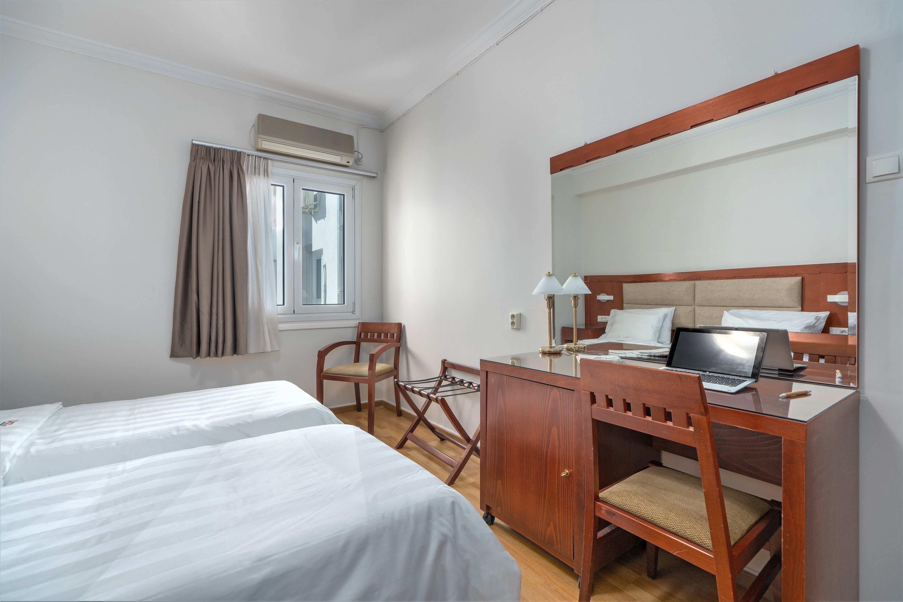 A hotel room with two beds, a desk with a laptop, a chair, a mirror, and a window with curtains.