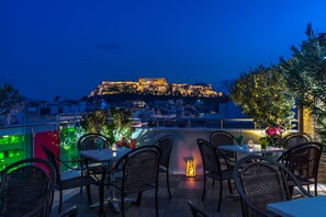 Un área de comedor al aire libre con sillas y mesas de mimbre, con vista a un edificio histórico con ruinas iluminadas.