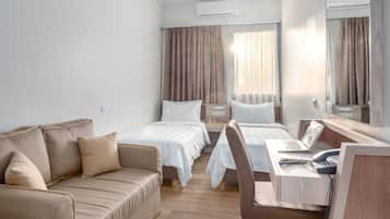 Ein Hotelzimmer mit Holzboden, einer Couch, zwei Einzelbetten, einem Schreibtisch mit einem Laptop, einem Stuhl, einem Spiegel und einer Klimaanlage.