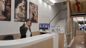 A reception desk with a woman on the phone, artwork on the wall, a TV, and a staircase. - Attalos Hotel (Athens)