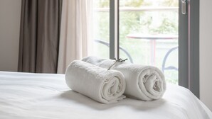 A neatly made bed with rolled white towels and a sprig of rosemary, in a room with a large window and curtains. - Attalos Hotel (Athens)