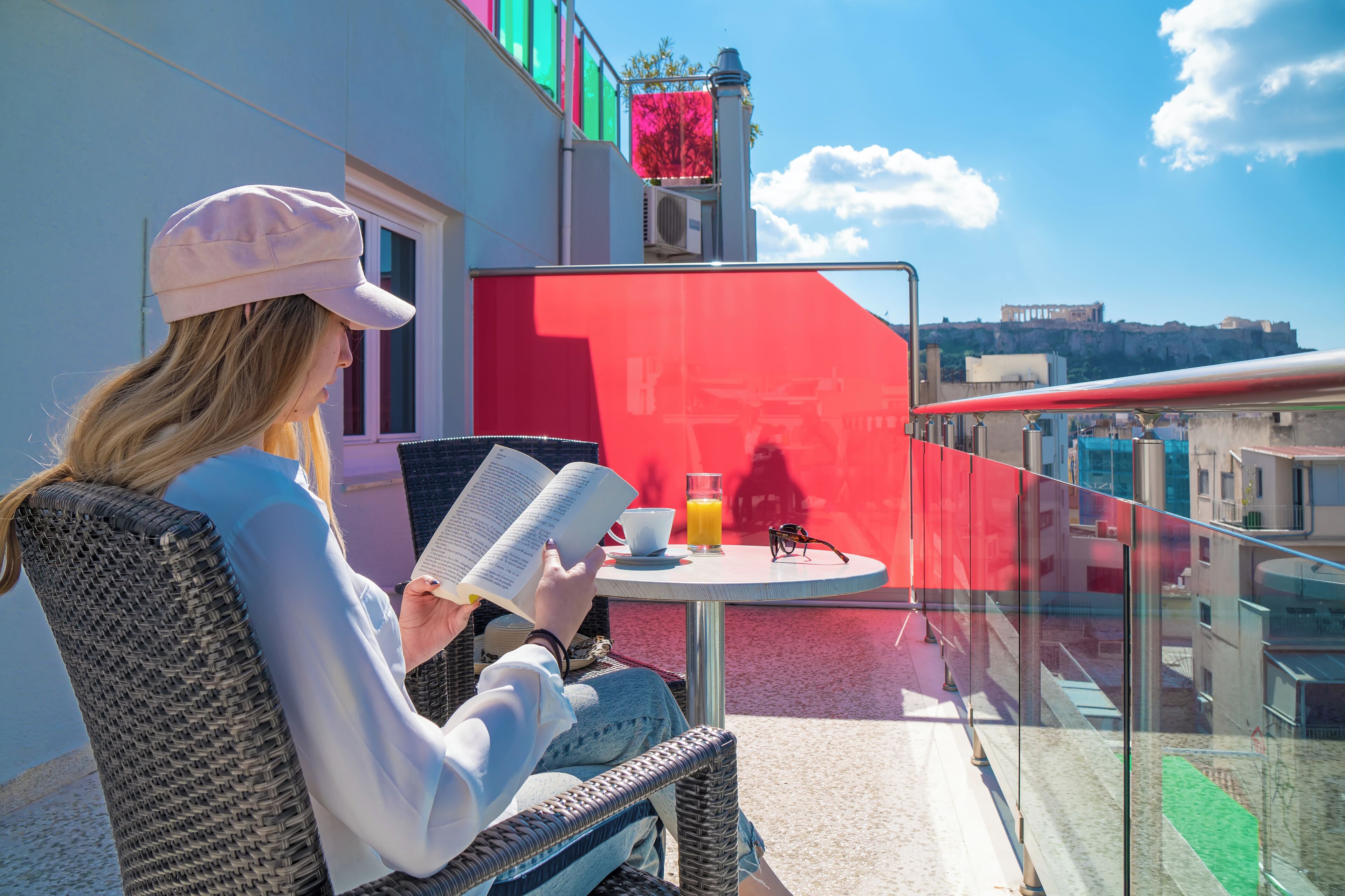 A person reading a book on a balcony with a table, a chair, and a glass of orange juice.