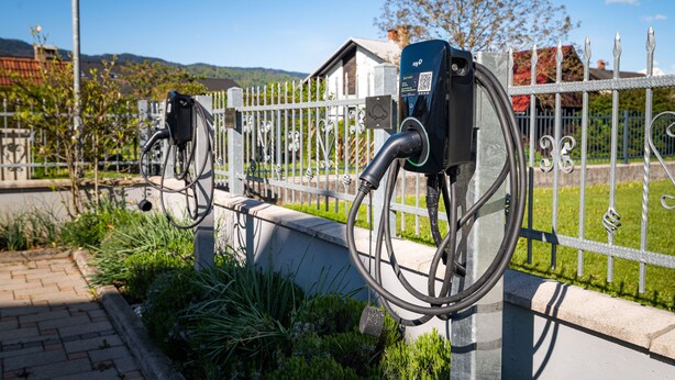 A bicycle charging station with a QR code, a metal fence, and a house in the background. - Penzion Kovac (Radovljica)