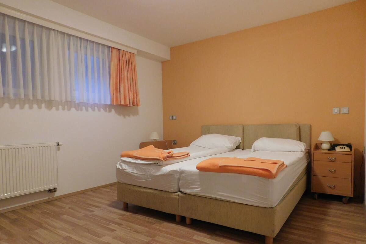 A double bed with white and orange bedding, a nightstand with a lamp, and a window with curtains.