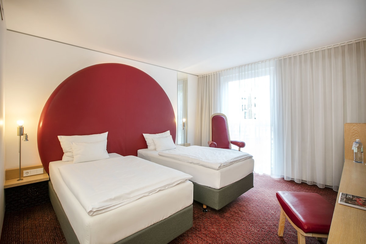 A hotel room with two beds, a red headboard, a red bench, a desk, and a mirror.