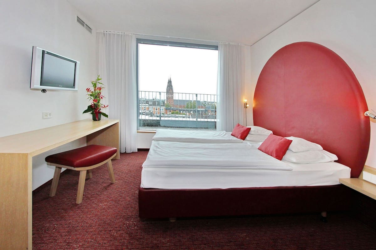 Una habitación de hotel con una cama, un escritorio, una silla, un televisor, una ventana con vistas a un edificio y un cabecero rojo.