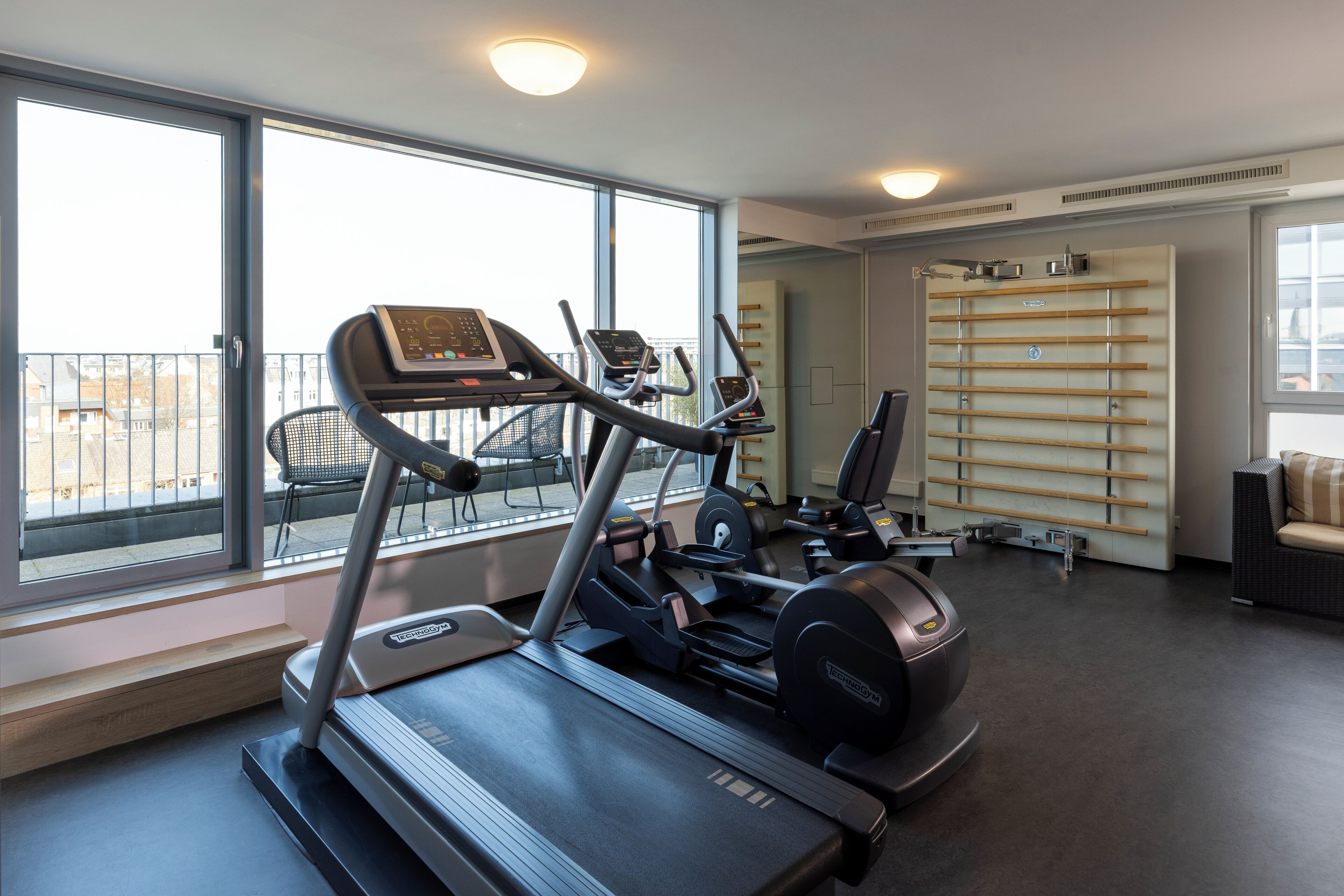 Uma academia com esteiras, um aparelho de exercícios fixo na parede e uma área de estar com vista para a cidade.