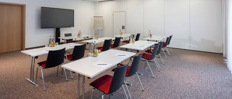A conference room with multiple tables, chairs, a large screen, and a carpeted floor.