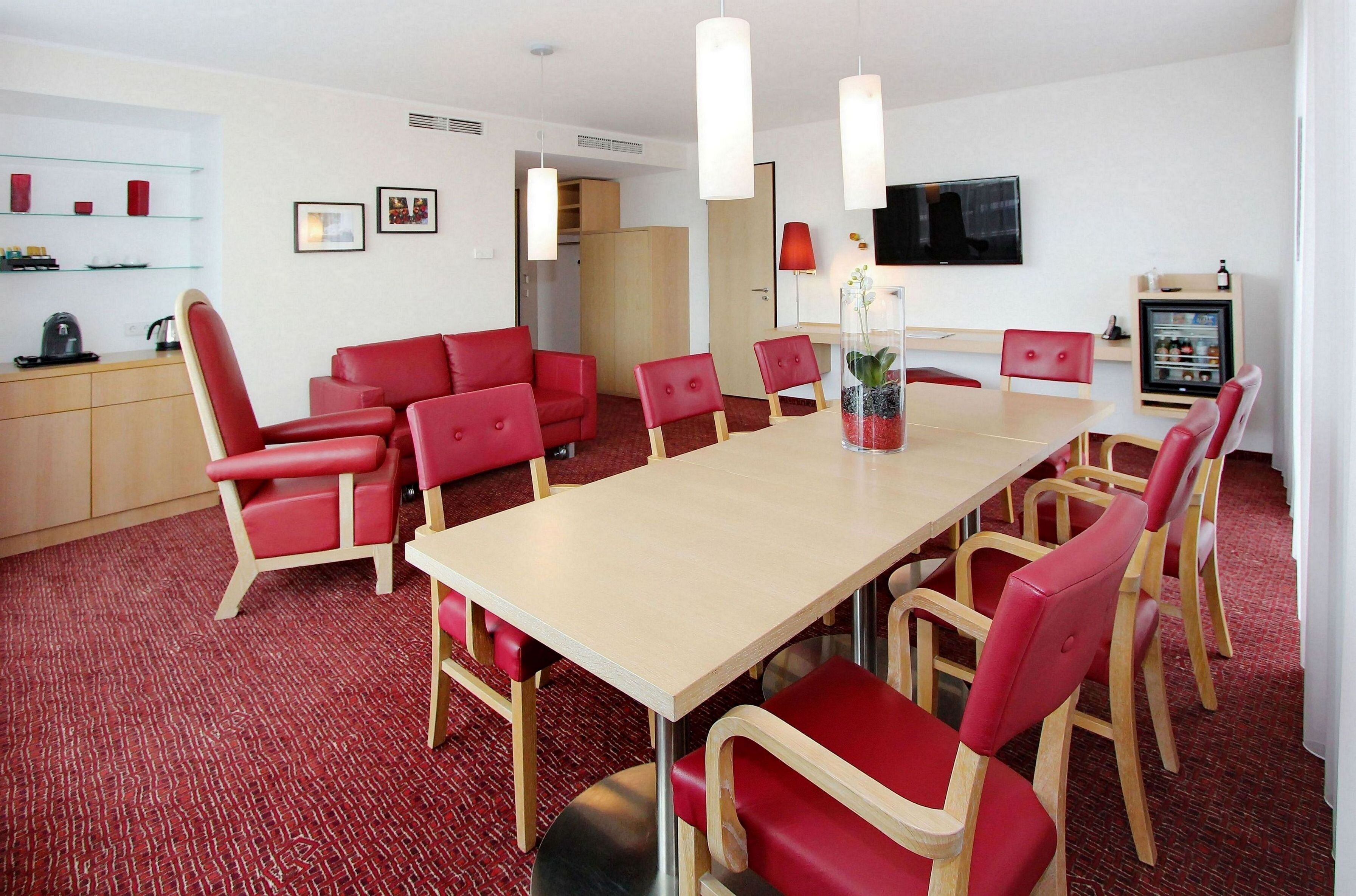 A modern dining area with a long table, red upholstered chairs, a television mounted on the wall, and a refrigerator with beverages.