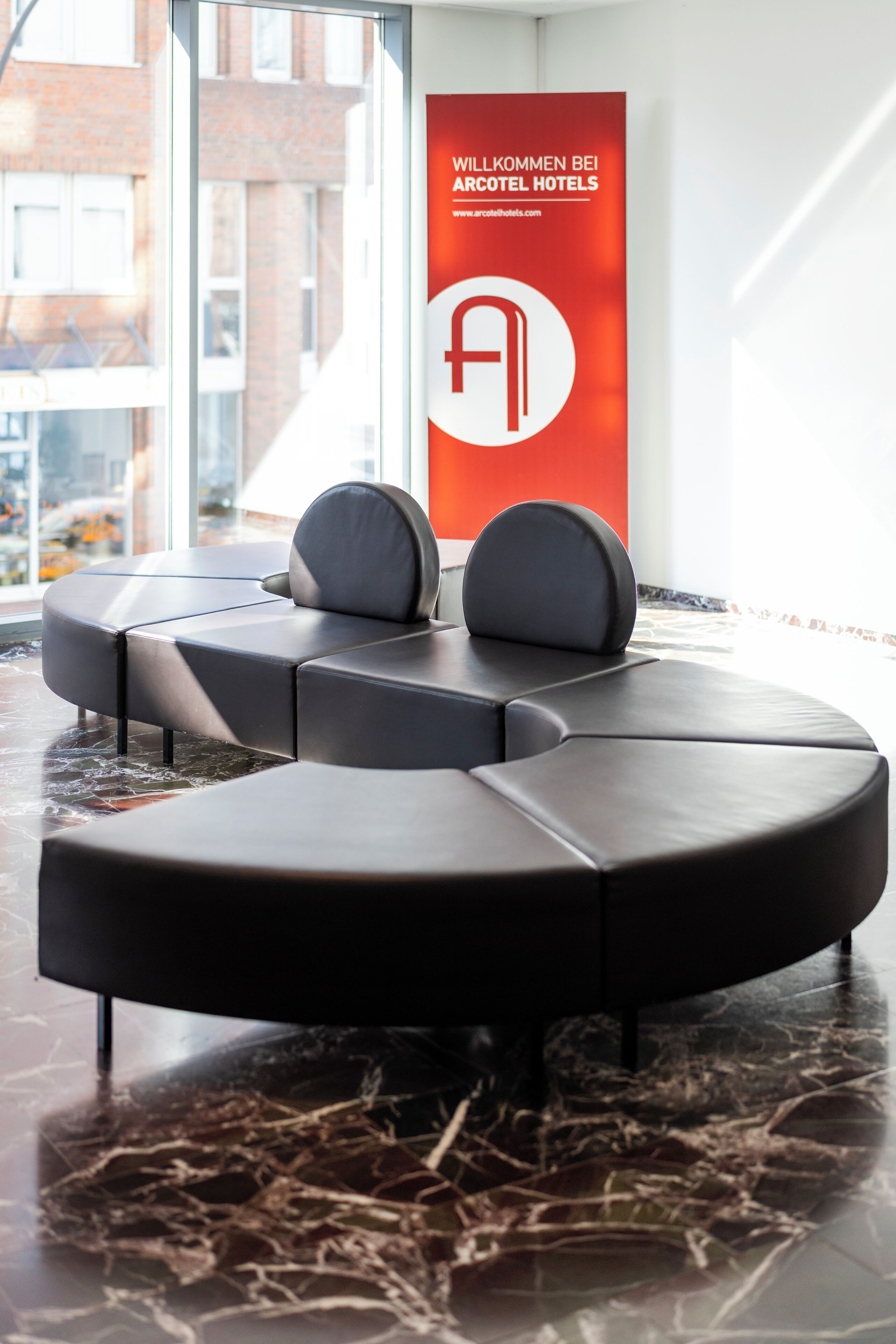 A modern hotel lobby with a sectional sofa, a large window, and a red banner with the hotel's logo.