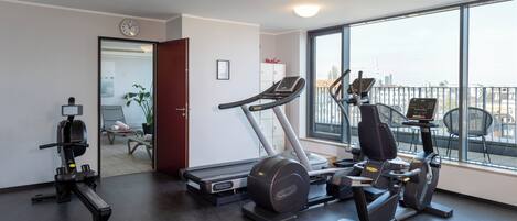 Een sportschool met loopband, elliptische trainer en krachtapparaat, grote ramen en uitzicht op de stad.