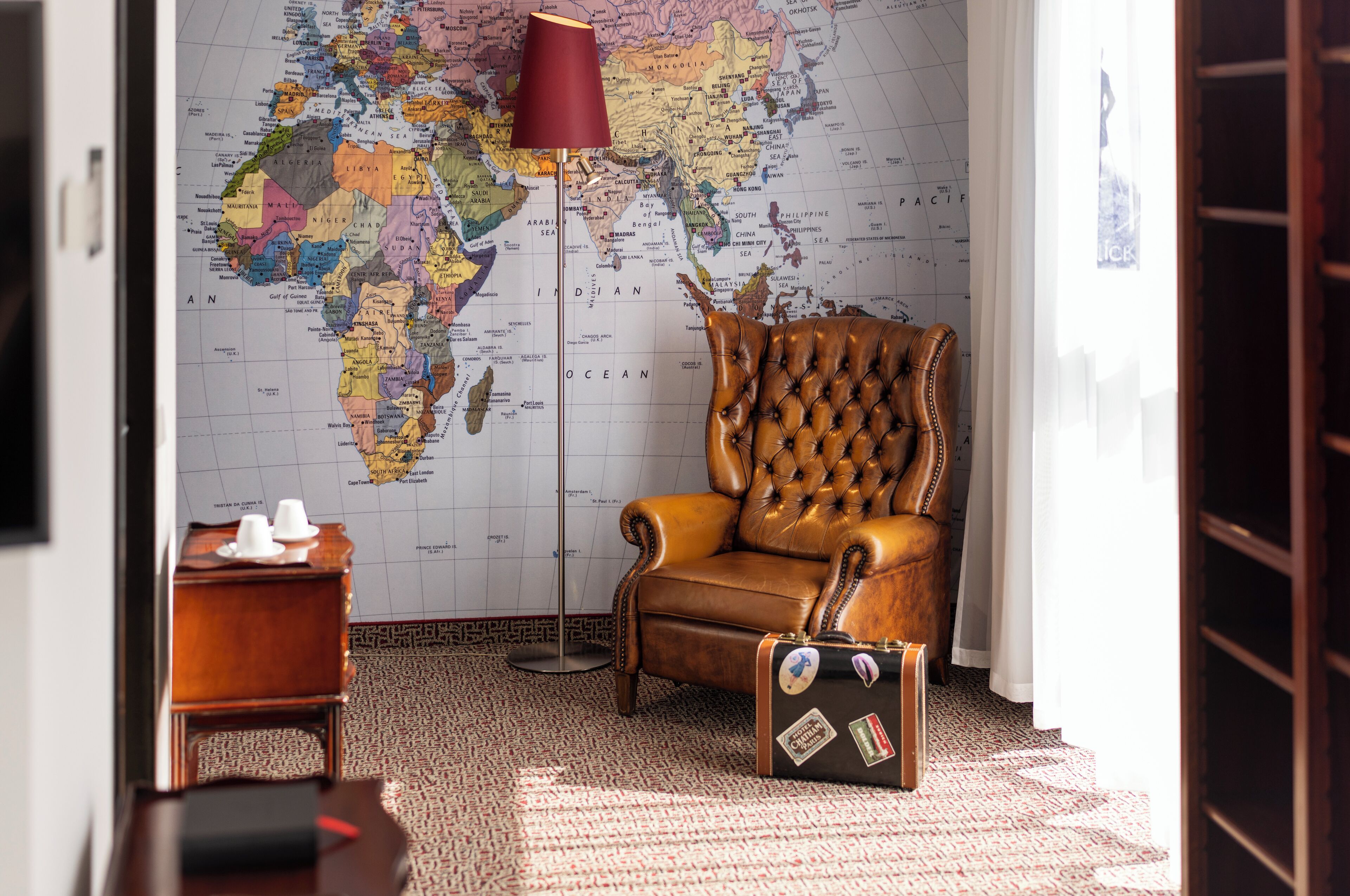 Une pièce avec une grande carte du monde accrochée au mur, un fauteuil en cuir, une petite table en bois avec des tasses et une valise ornée d’autocollants de voyage.
