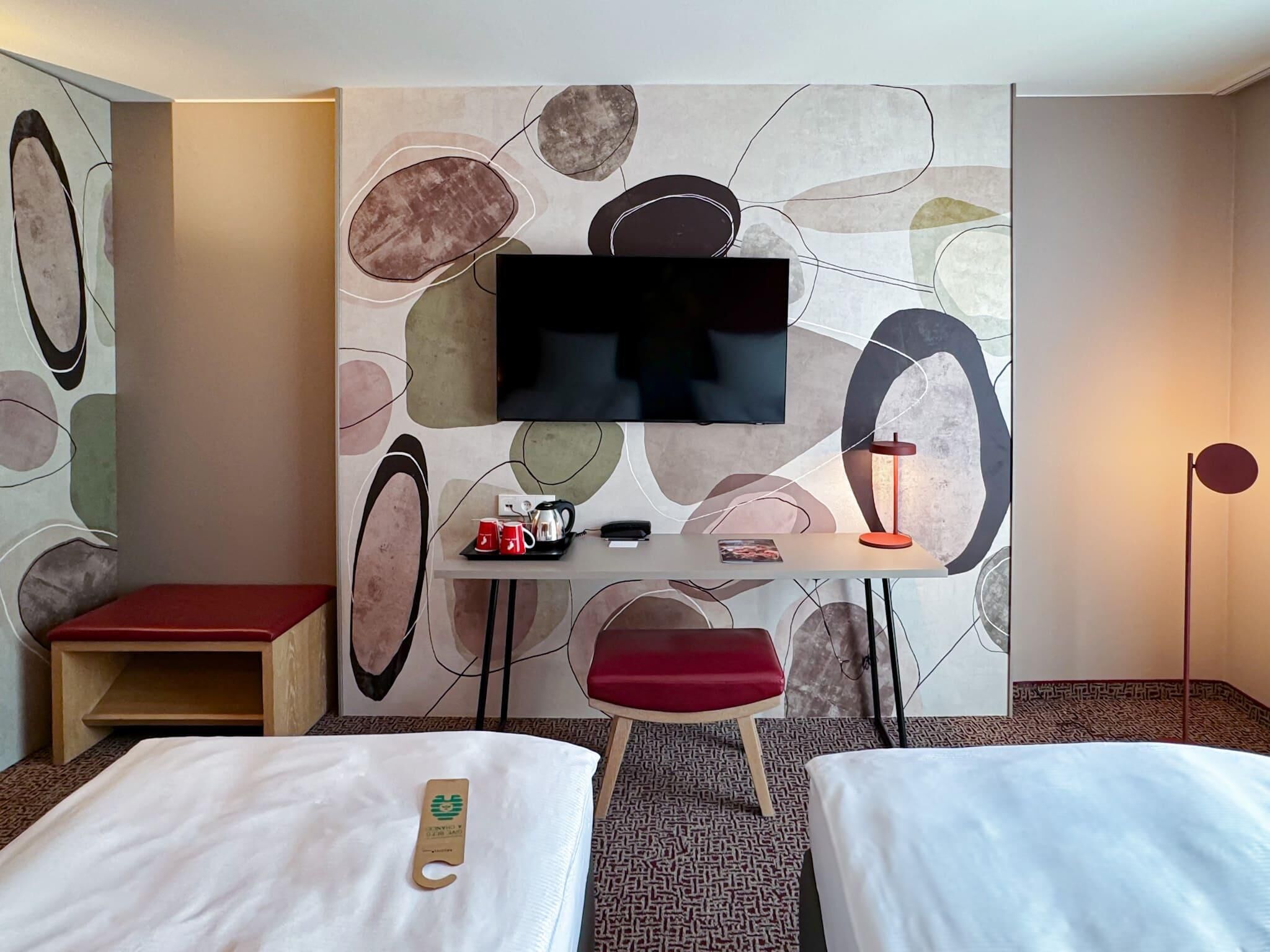 Habitación de hotel con dos camas, un televisor montado en la pared, un escritorio pequeño con una tetera, una taza roja y una silla roja.
