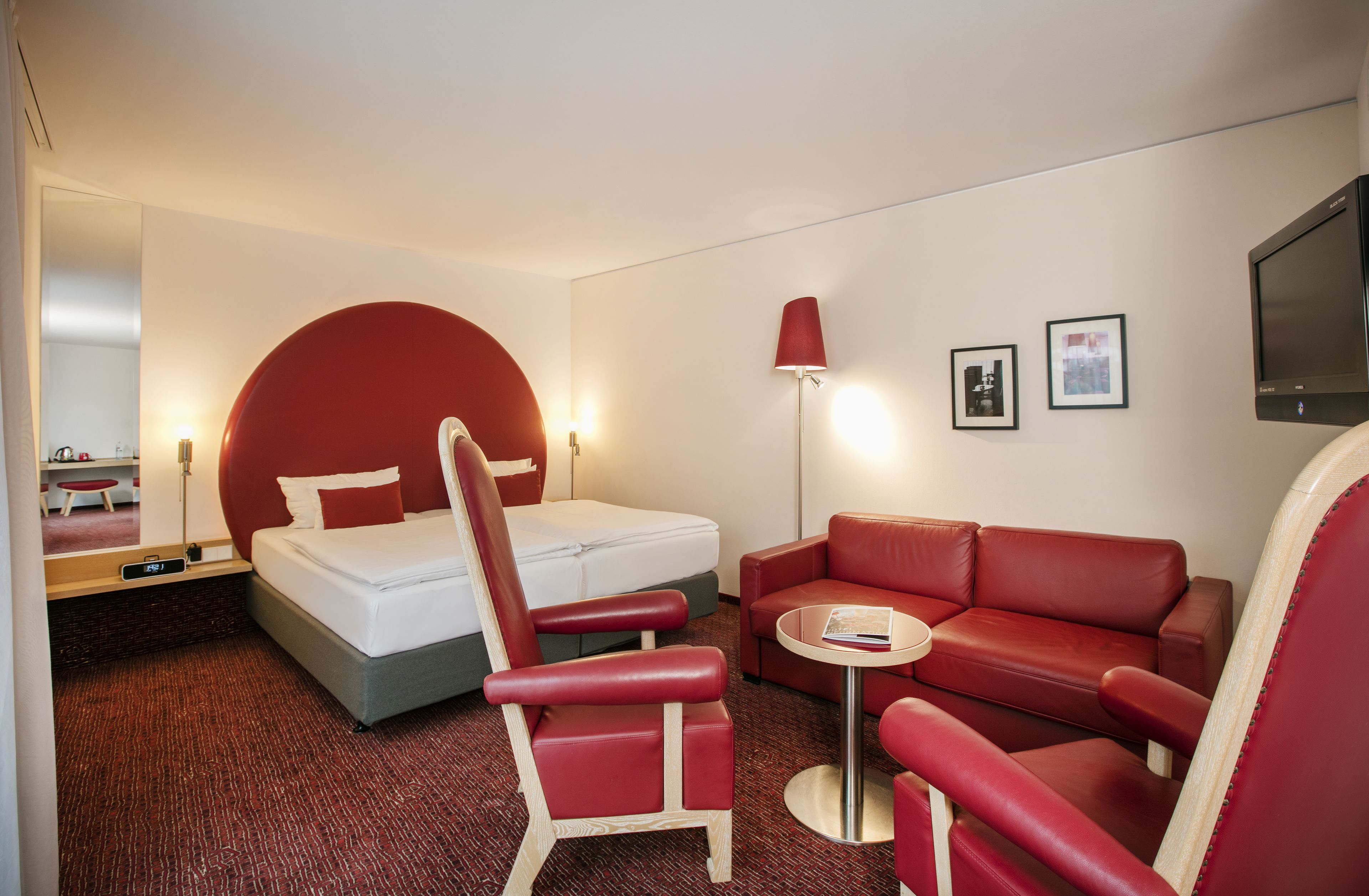 Et hotelværelse med en seng, en rød lænestol, en rød sofa, et lille bord og et fjernsyn.
