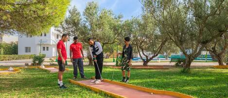 A group of people playing croquet in a park with trees and a tennis court in the background.