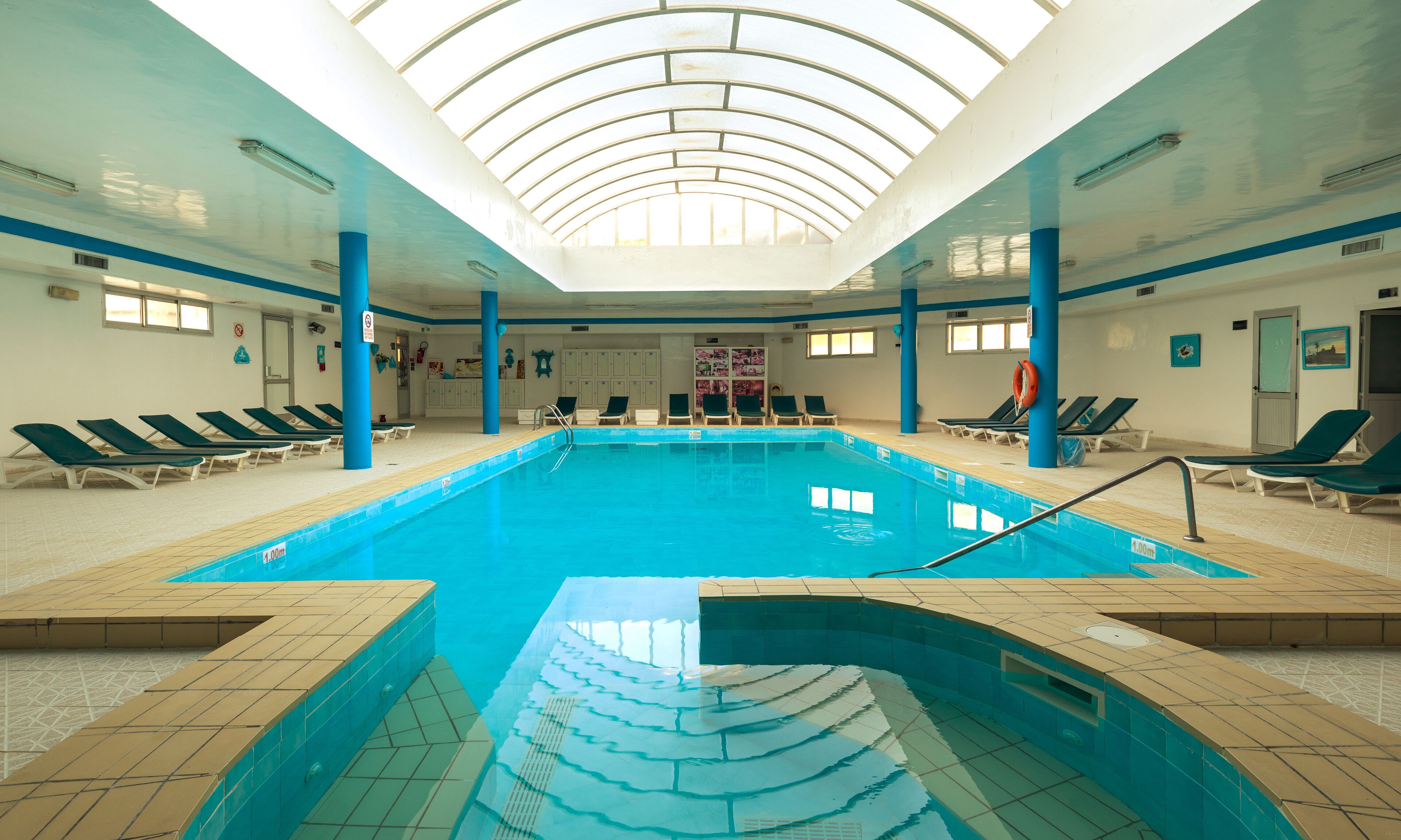 Une piscine intérieure à l’eau d’un bleu cristallin, entourée de chaises longues et dotée d’un sol carrelé.