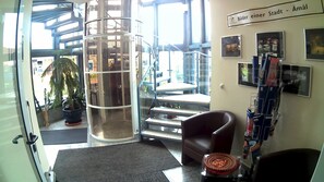 A lobby with a glass elevator, a staircase, a brown leather chair, a small table with a decorative item, and a wall with framed pictures and a sign. - Hotel Christinenhof Gadebusch (Gadebusch)