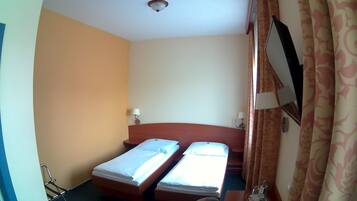 A small hotel room with two single beds, a wooden headboard, a bedside table with a lamp, and a window with curtains.