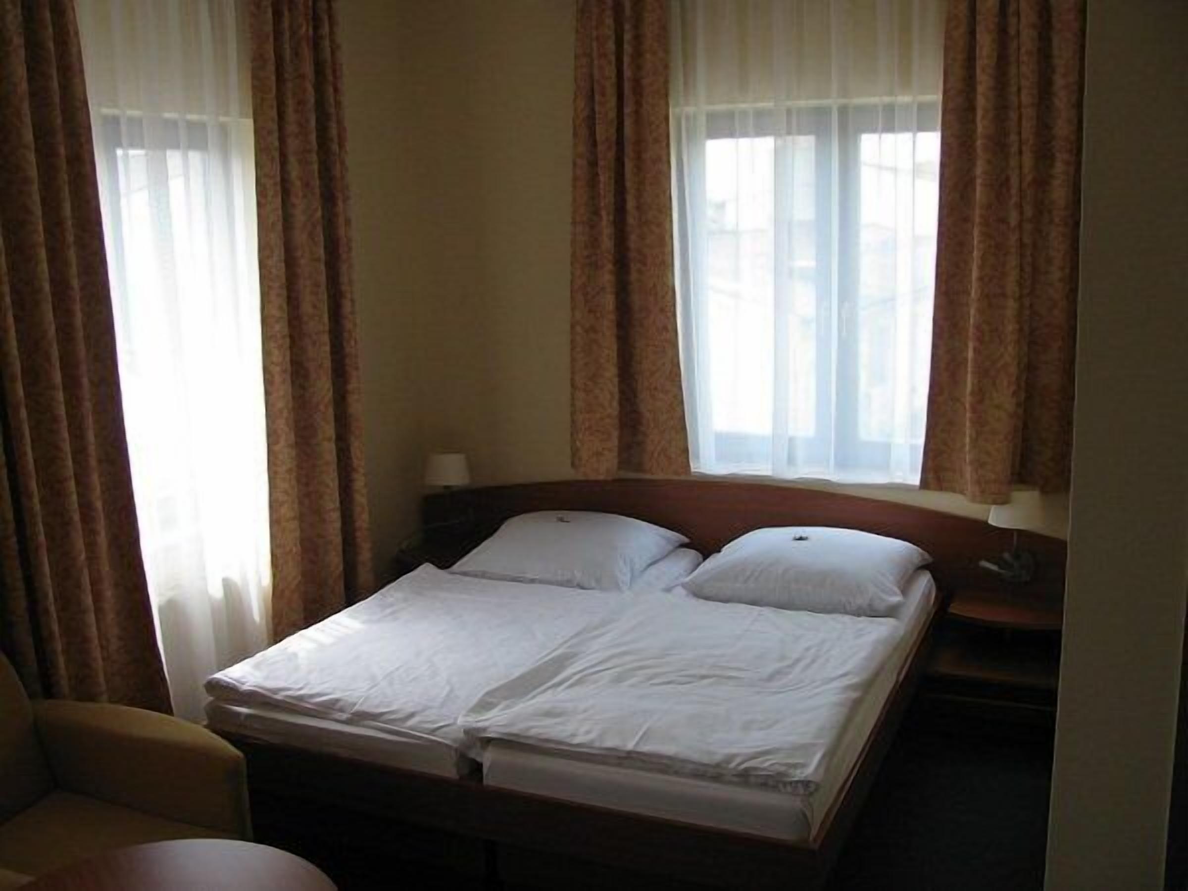 Quarto de hotel com uma cama, duas almofadas, mesa de cabeceira, abajur e janela com cortinas.
