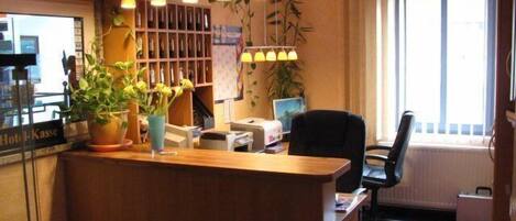 Un comptoir d’accueil d’hôtel avec un comptoir en bois, un ordinateur et un fauteuil de bureau noir. Des plantes en pot sont posées sur le comptoir, et une fenêtre avec des rideaux se trouve à l’arrière-plan.