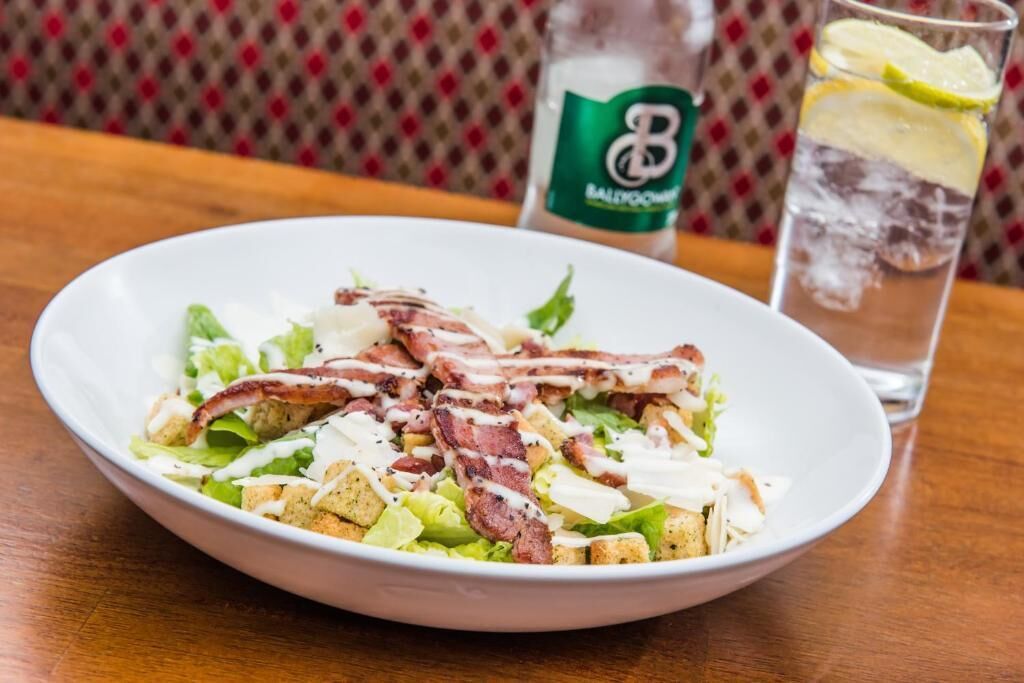 Un bol de salade avec des lardons et des croûtons, un verre d’eau avec une tranche de citron vert et une bouteille d’eau Balducci sur une table en bois.