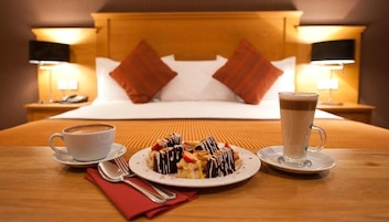 Una mesa de desayuno ordenada con un plato de postre, una taza de café y un vaso de bebida caliente en una habitación de hotel.