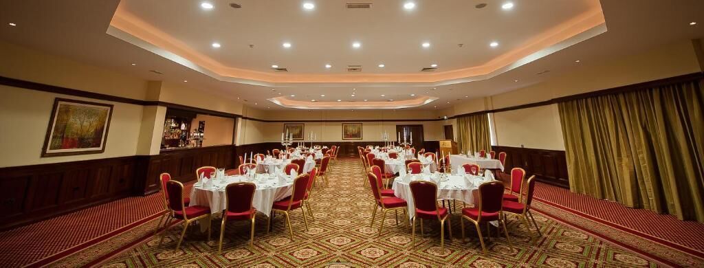 Un salón de banquetes con mesas redondas y sillas rojas, suelo alfombrado y un techo alto con iluminación empotrada.